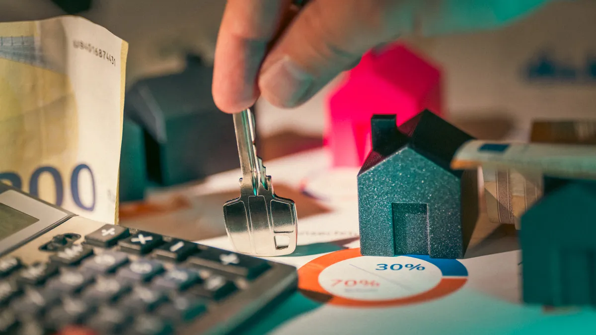 Hand holding a key beside a house model, calculator, and chart